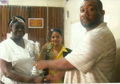 Long serving Secretary/CEO of the Rose Hall Town Youth and Sports Club, Hilbert Foster (right), hands over the trophy and medallions to Deputy Principal of New Amsterdam Secondary School, Shondell Phillips, while another member of staff displays some more medallions at center.