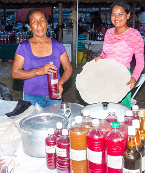 Lynette Adrian holds a bottle of her special ‘Fly’, while her niece, Nadia, shows the huge cassava bread they sell with their wild meat dishes