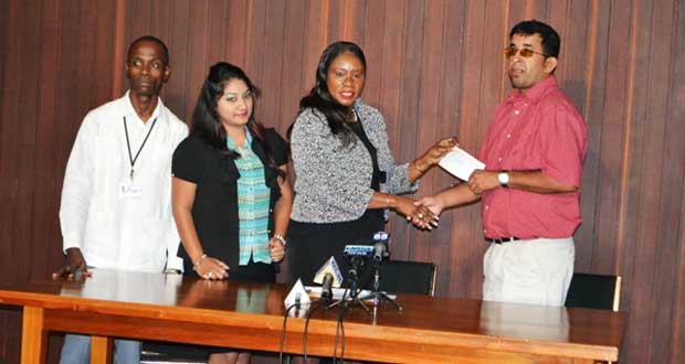 Minister of Human Services and Social Security, Ms. Jennifer Webster handing over the cheque to Public Relations Officer for the Council of the Organisation of People with Disability and member of the Board of Governors of the Guyana Society for the Blind, Ganesh Singh, while staff members from the OLPF Secretariat look on
