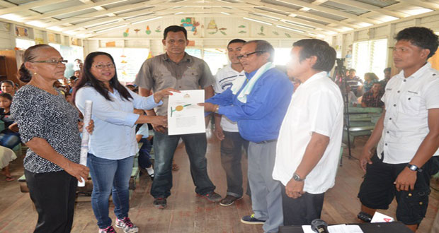 Minister of Amerindian Affairs, Ms. Pauline Sukhai handing over the title document to Toshao of Kariako, Mr. Lewis Samuels in the presence of Liaison to the Ministry, Ms. Yvonne Pearson, and residents of Kariako