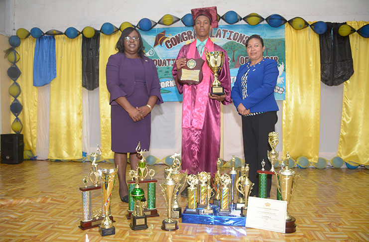 Valedictorian Mark Ryan Wilburg standing between Minister of Health within the Ministry of Health, Dr.  Karen Cummings Minister of Public Affairs in the Office of the President, Ms. Dawn Hastings-Williams, showing off some of the trophies and other gifts he received