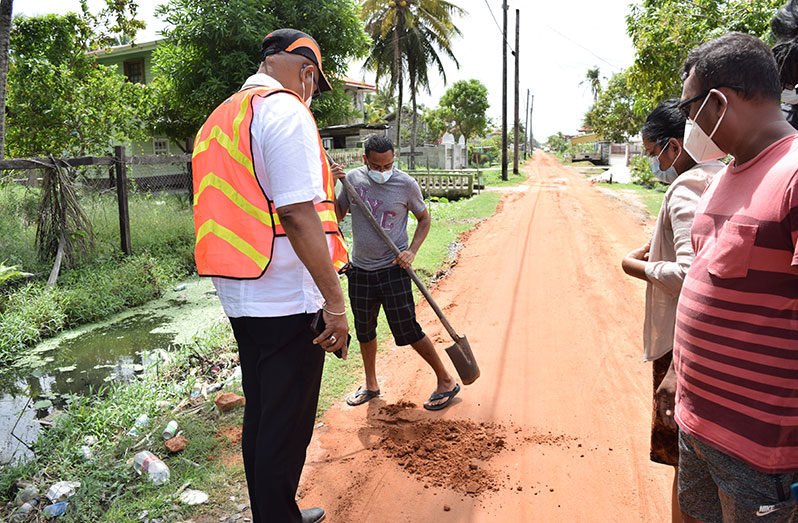 Minister Edghill watches on as a resident digs a section of the road, in Number Two Village, to ensure it meets the required thickness