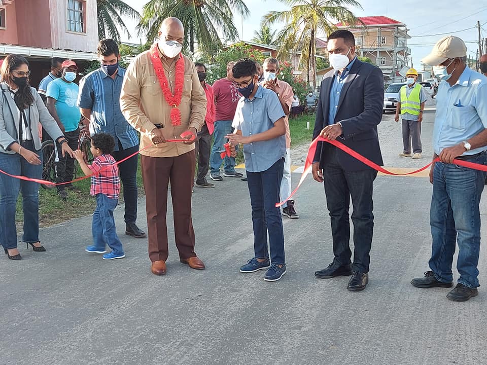 Minister of Public Works Juan Edghill and Minister of Local Government and Regional Development, Nigel Dharamlall, assist a resident of Good Hope in cutting the ceremonial ribbon to officially commission the upgraded phase two road network, on Thursday