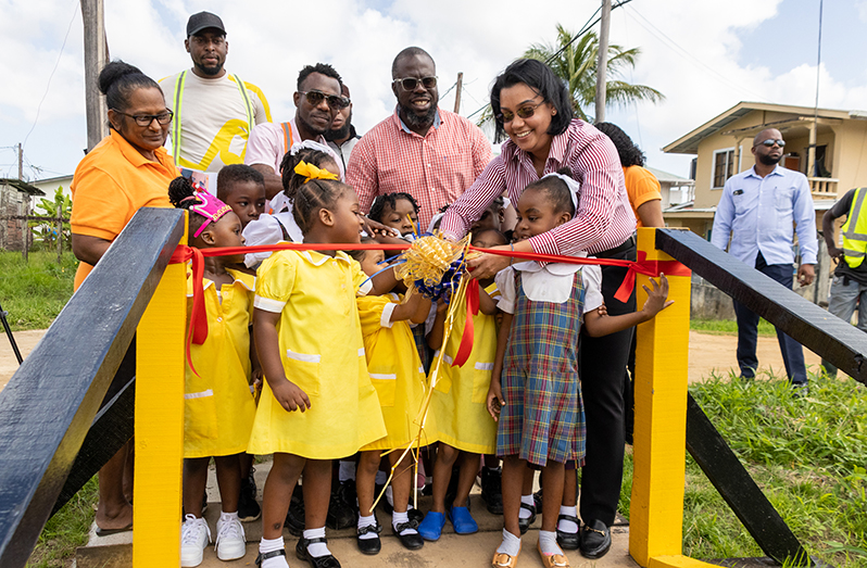 Minister within the Ministry of Housing and Water, Susan Rodrigues, assists children in cutting the ribbon to commission one of three newly constructed timber bridges leading to the school