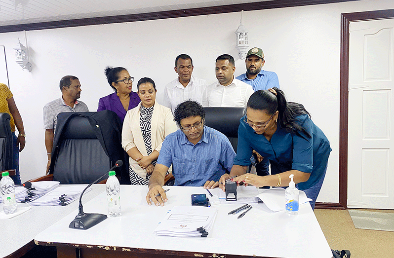 A contractor signing in presence of Vladim Persaud, Permanent Secretary of the Ministry of Public Works, as regional officials and Minister within the Public Works Ministry, Deodat Indar look on