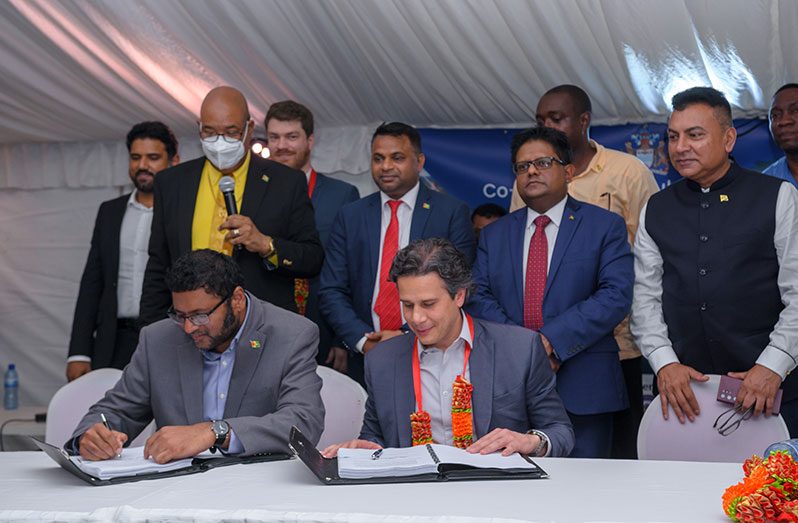Permanent Secretary of the Ministry of Public Works, Vladim Persaud, along with a representative of the Quieroz Galvao, signs the contract for the upgrade of the Linden to Mabura Hill Road in the presence of Public Works Minister Juan Edghill (standing second from left); Minister within the Ministry of Public Works, Deodat Indar (fourth from left); Finance Minister, Dr Ashni Singh (second from right); and Director of Go-Invest, Dr Peter Ramsaroop (first from right) (Delano Williams photo)