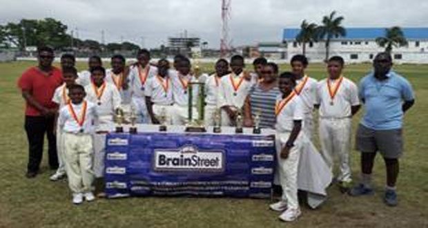 Champs again! The victorious DCC Under-15 team strike a pose with the GCA/BrainStreet Under-15 banner and the trophies they won during the fifth staging of the competition which culminated last Sunday at the Malteenoes Sports Club ground. At right is coach Garvin Nedd.
