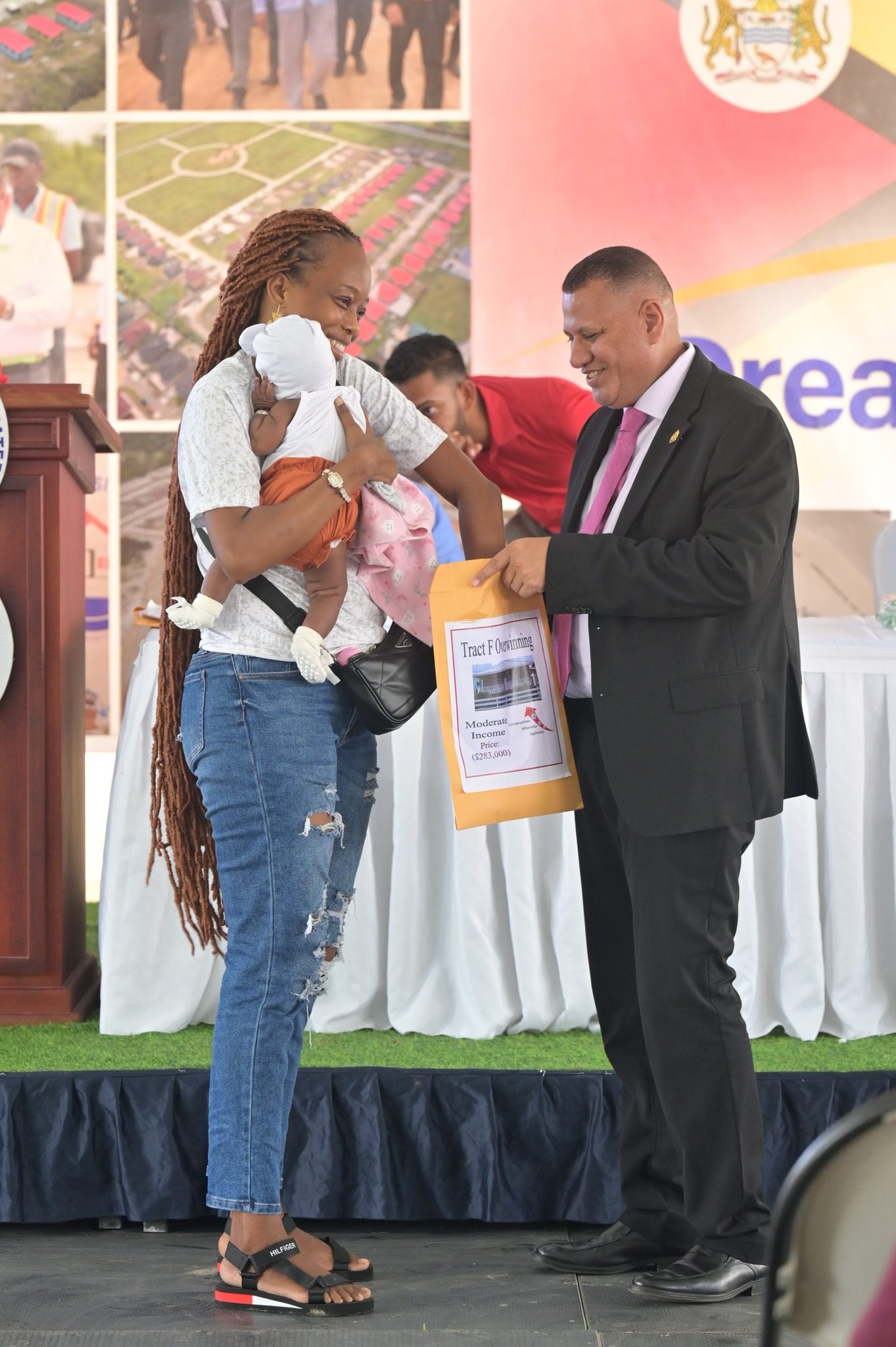 A mother holding her child pulls her lot number from an envelope being held by Minister of Housing and Water, Collin Croal, at the two-day 'Dream Realised' Housing Drive at the State House, Region Six