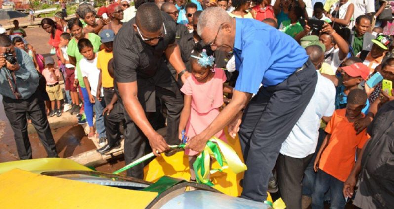 A Linden girl is assisted to cut the ribbon with President David Granger to commission a boat for use of children in the Demerara River. At the ceremony, the President announced that Books and Breakfast will be added to his Bus, Bicycles and Boats initiative, to ensure that every child gets to school.