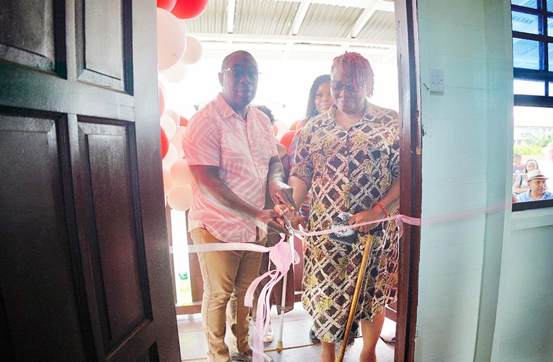 Minister within the Office of the Prime Minister with Responsibility for Public Affairs, Kwame McCoy, assists Nicole Fraser in cutting the ribbon to signify the handing over of her new residence