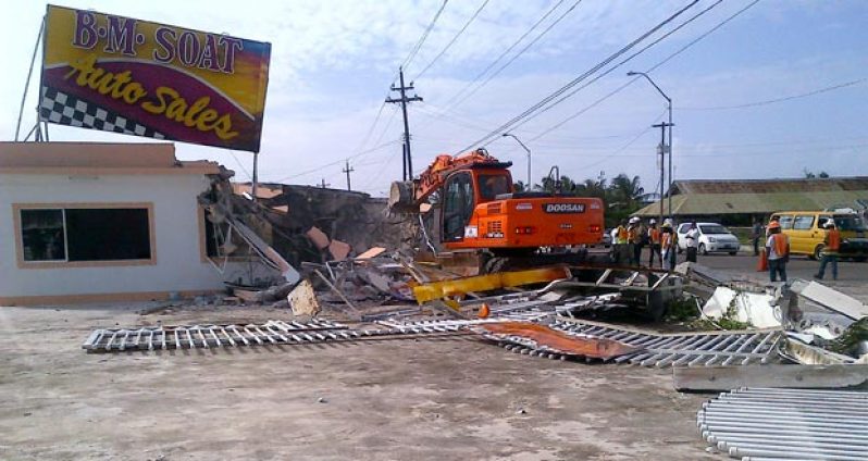 Photo shows  a broken-down fence in the foreground while the excavator works to bring down the office of the auto sale company yesterday.