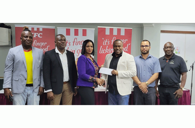 KFC Guyana Marketing Manager, Pamella Manasseh, hands over the sponsorship to Co-Director of the Petra Organisation, Troy Mendonca, in the presence of Assistant Director of Sport Franklin Wilson (second from left), Nicholas Fraser (second from right) of the Allied Arts Division, Ministry of Education, Competitions Chairman at the Guyana Football Federation, Troy Peters (far right)  and Referee Coordinator, Wayne ‘Harry’ Griffith (far left)