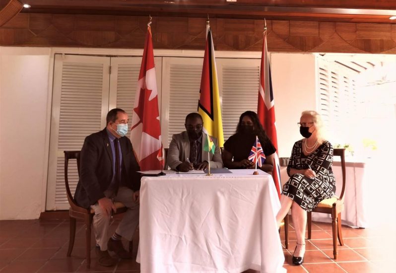 Minister Kwame McCoy signs the agreement as High Commissioner, Mark Berman (left), Nazima Raghubir and High Commissioner, Jane Miller (right) look on