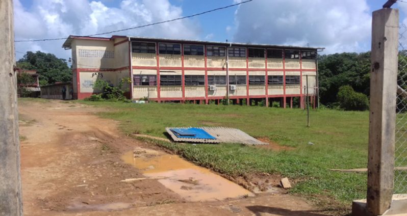 The Port Kaituma Secondary School in the background with a pool of water and part of the gate to the school in the foreground