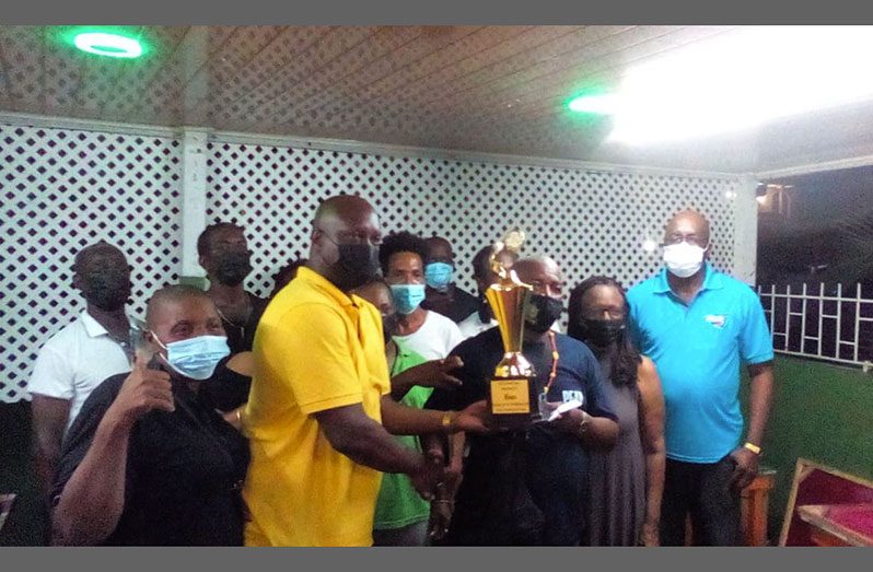 President of the Georgetown Dominoes Association Lyndon Boston hands over the winning trophy to the MVP and player Dexter Thompson of Spartans while team members and executives look on approvingly.