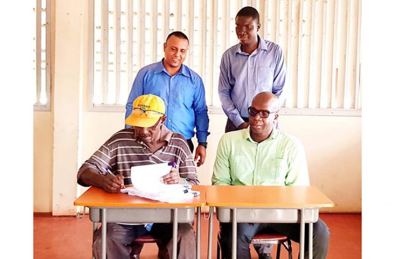 CCSP Project Manager, Clement Henry; Chairman of Bush Lot/ Adventure NDC, Colin Moore; and CCSP Procurement Officer, Mark Pertab look on as contractor Brindsley Ross signs the contact at the Kildonan Multi-Purpose Centre
