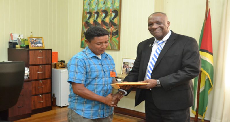 Minister of State Joseph Harmon hands over the money to Toshao of Toka Village, Delano Davis (Ministry of the Presidency)