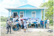 house_ Minister McCoy addresses the small gathering outside the new house which was built through the MoM programme. Also pictured sitting on a chair is the recipient, 80-year-old Dalia Lewis (MoM photo)