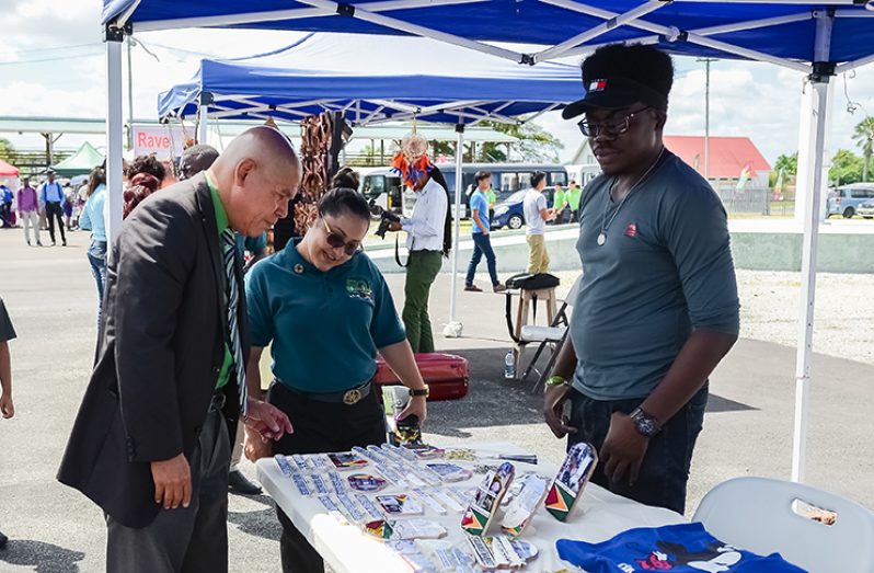 Minister of Social Cohesion, Dr. George Norton (left) checks out some of the products on display at Jamal Farley’s (right) booth, as Youth Village Coordinator, Adeti DeJesus looks on  (Delano Williams photos)